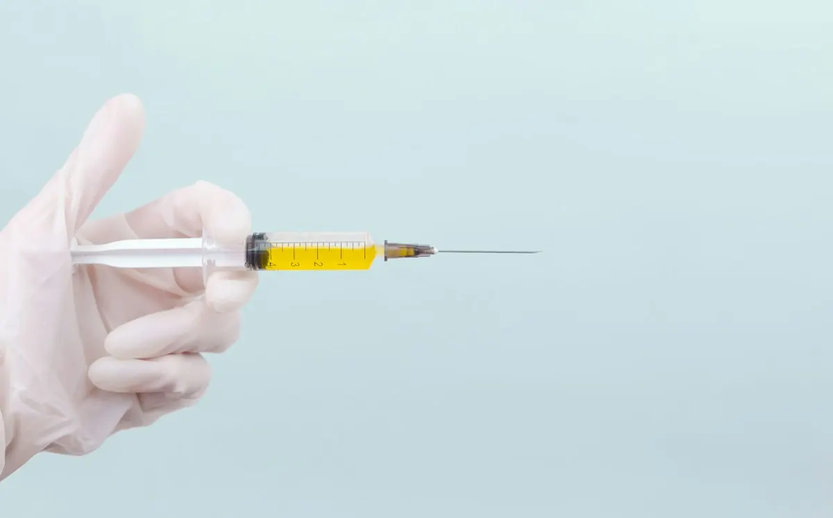 Mano con guante sosteniendo una jeringa con medicamento, fondo celeste