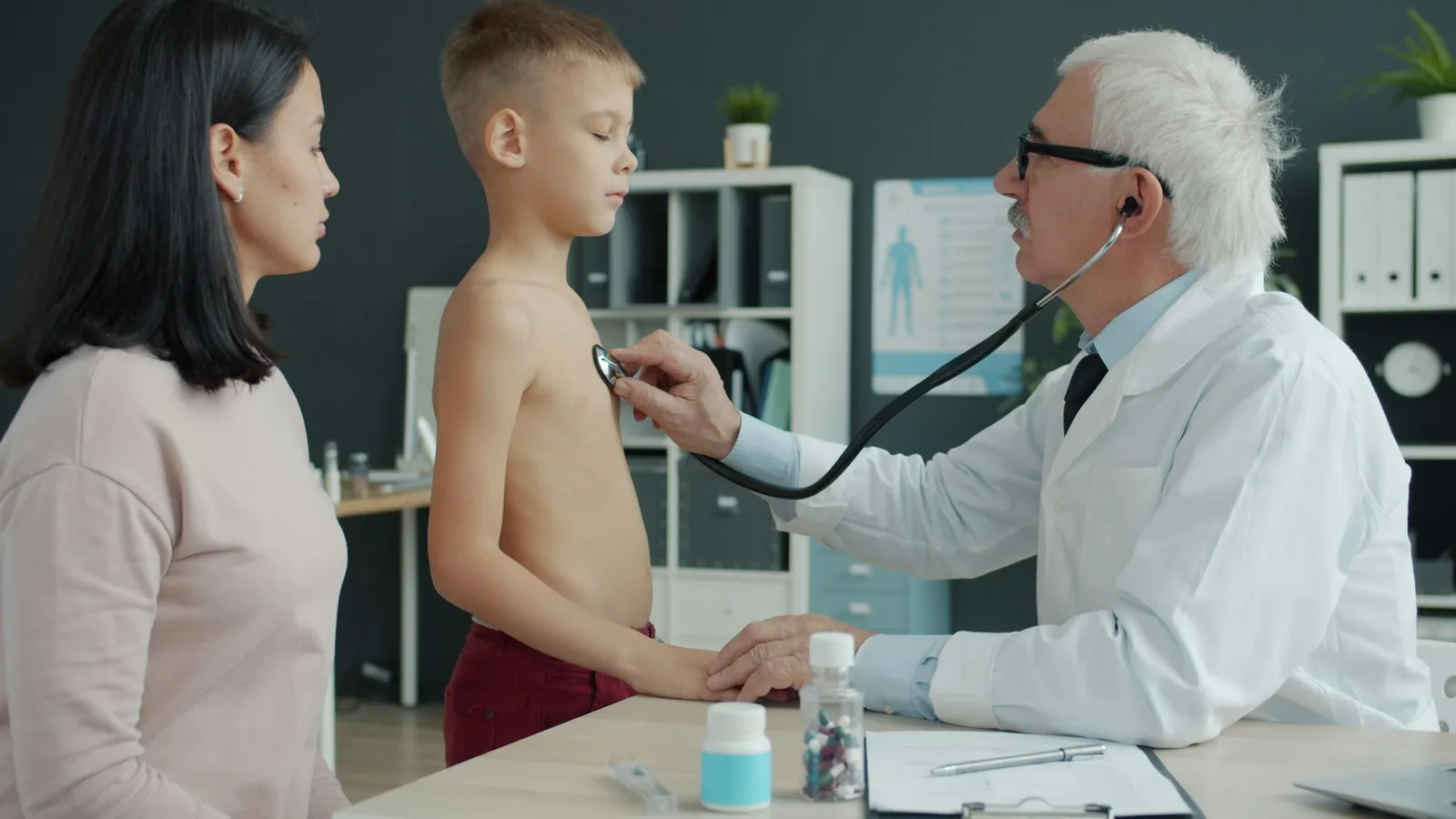 Médico mayor escuchando el pecho de un niño con un estetoscopio mientras su madre lo acompaña